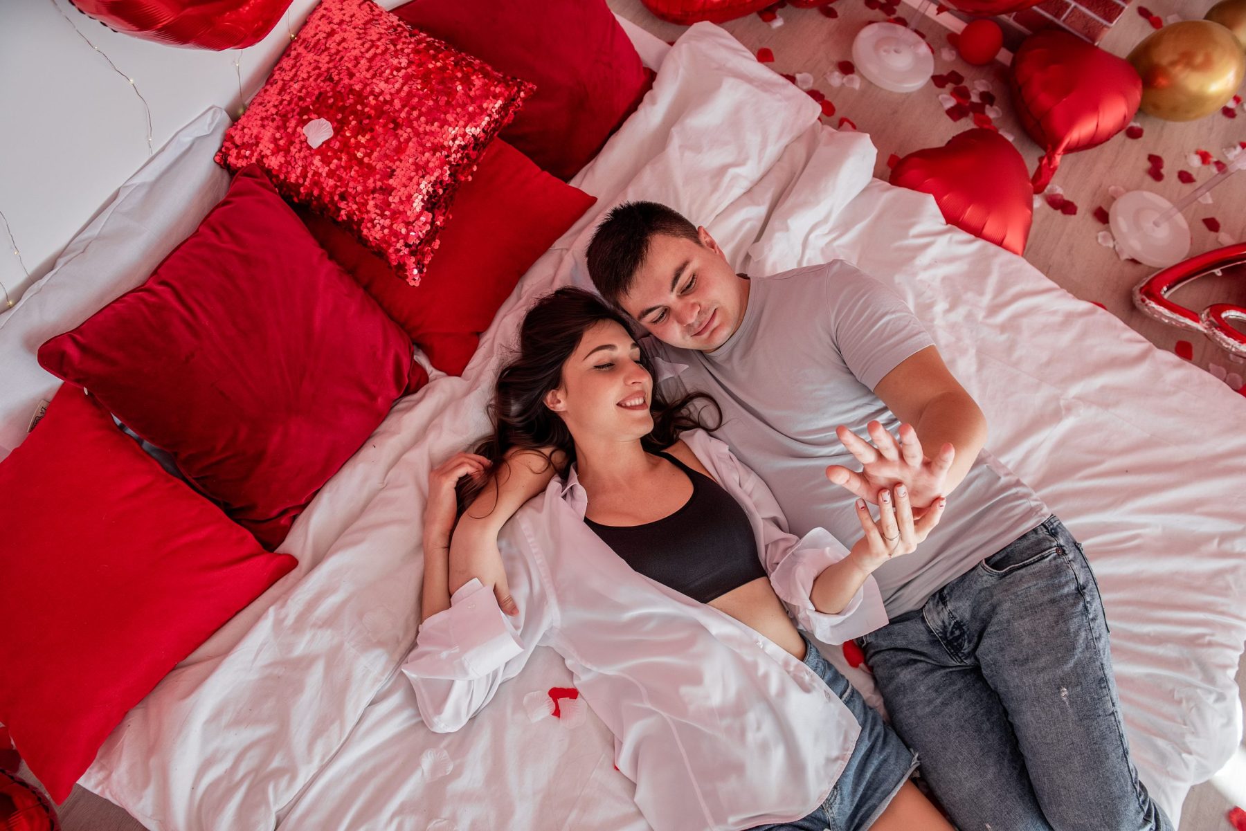 Vista cenital de pareja abrazada sobre cama blanca con globos rojos en forma de corazón, símbolo del Nakshatra Pūrva Phalgunī en la astrología védica